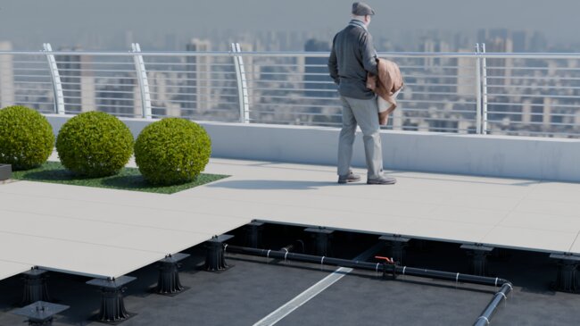 Cutaway view of an elevated rooftop terrace with a man standing near the railing, revealing the adjustable pedestal system, drainage, and piping underneath the floor tiles.