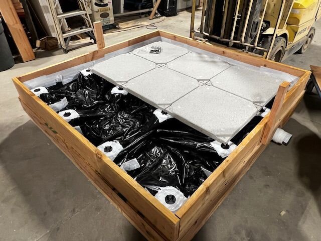 Large wooden pedestal frame with black plastic liner and light grey porcelain pavers being installed for a rooftop deck setup