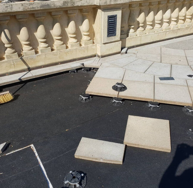 Installation of outdoor stone pavers on adjustable plastic pedestals over a black waterproof roof membrane.