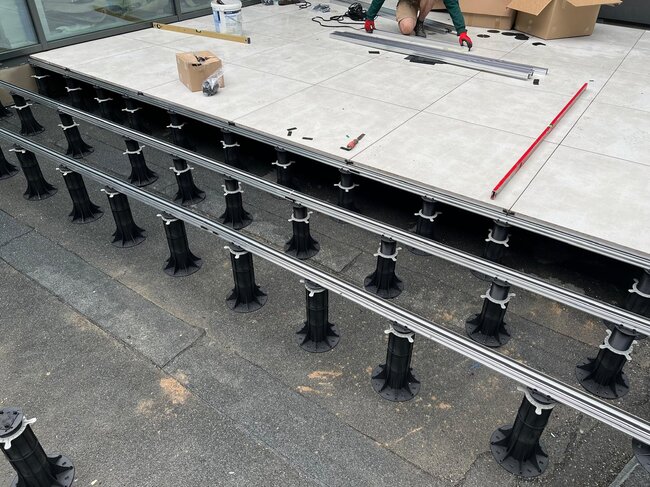 Installation of an outdoor raised patio using adjustable plastic pedestals and aluminum support joists with ceramic paving tiles.