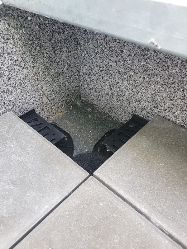 Corner placement of trimmed paving pedestals supporting raised outdoor floor tiles against a textured wall.