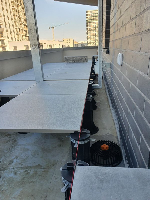 Red laser level line aligning porcelain tiles on tall adjustable pedestal supports along a building wall.