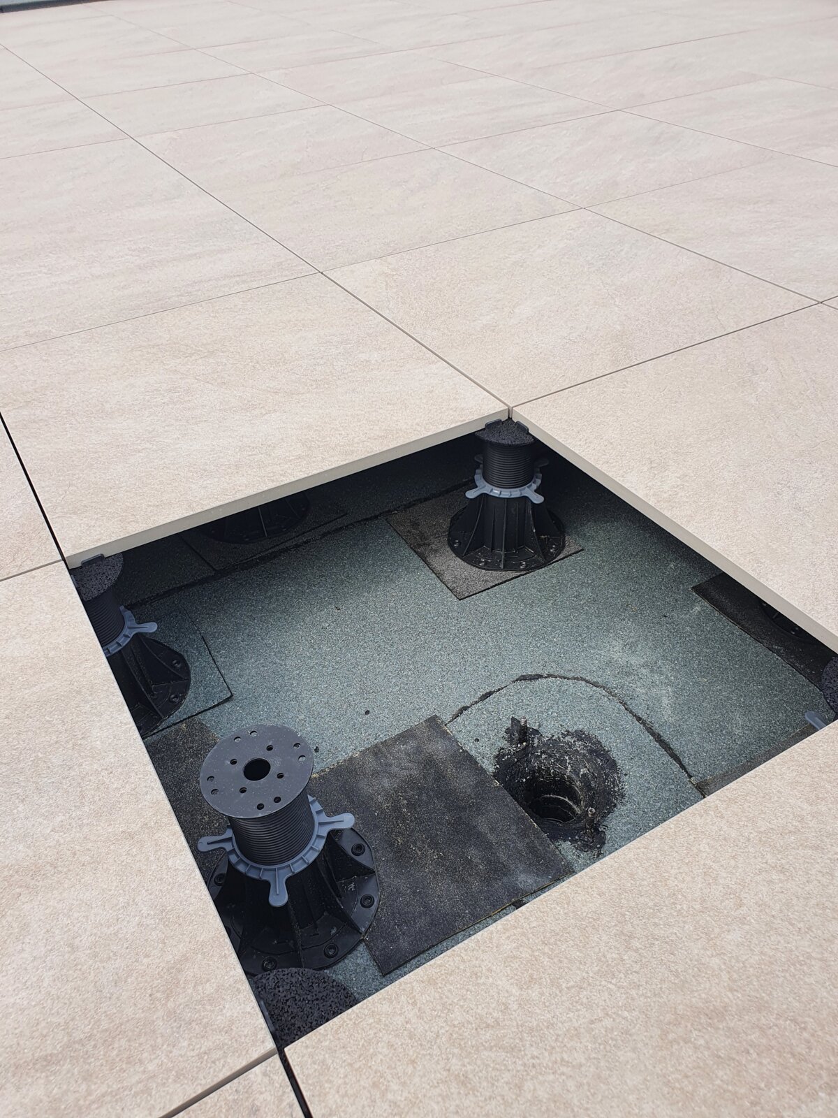 Top-down view of an open terrace paver section revealing an adjustable pedestal support and roof drain underneath.
