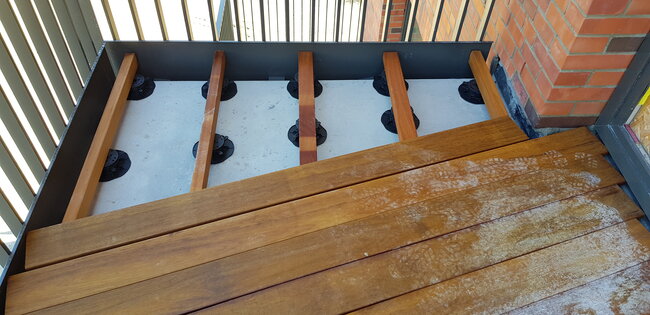 Balcony decking installation showing wooden joists supported on adjustable pedestal risers over a concrete base.