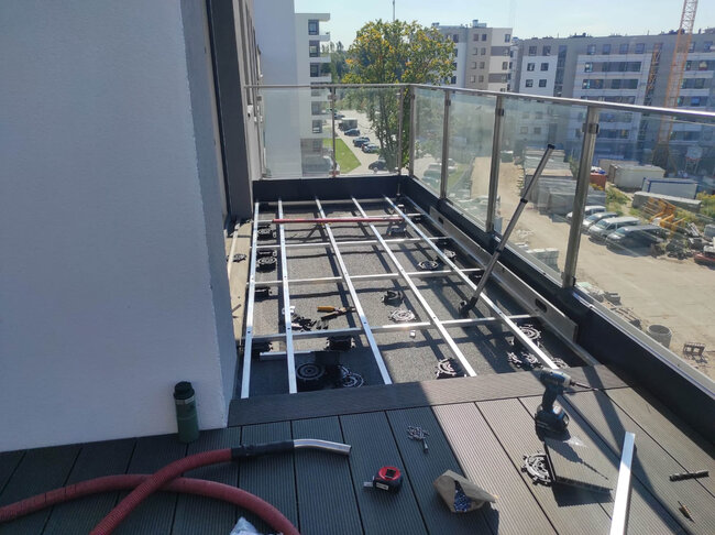 Balcony decking installation in progress showing aluminum joist framing, adjustable pedestals, and composite deck boards.