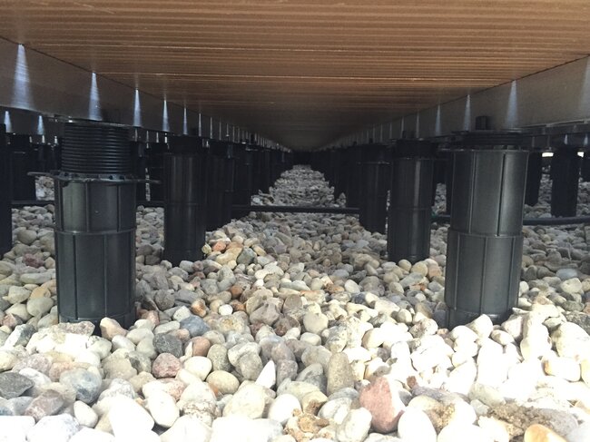 Adjustable pedestal supports installed under wooden decking over gravel.