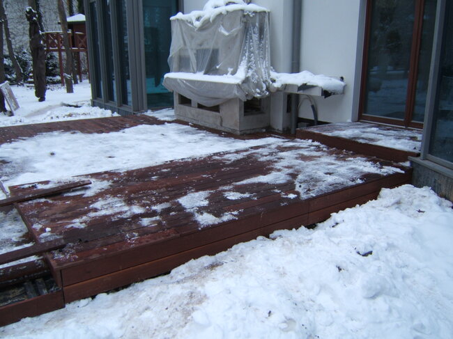 A dark wooden terrace deck partially covered with melting snow outside a house in winter.