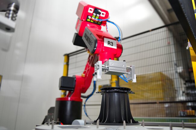 Red robotic arm assembling a black adjustable paving pedestal in an automated factory.